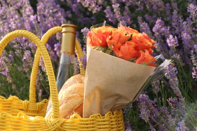 Photo of Yellow wicker bag with beautiful roses, bottle of wine and baguettes in lavender field, closeup