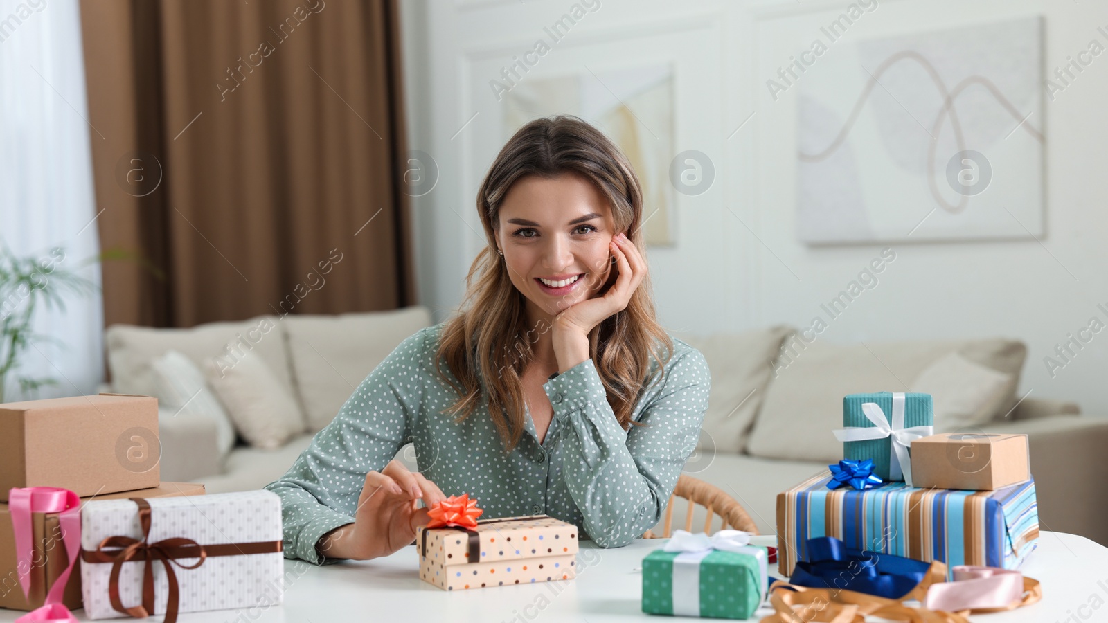 Photo of Beautiful young woman decorating gift box with bow at table in living room