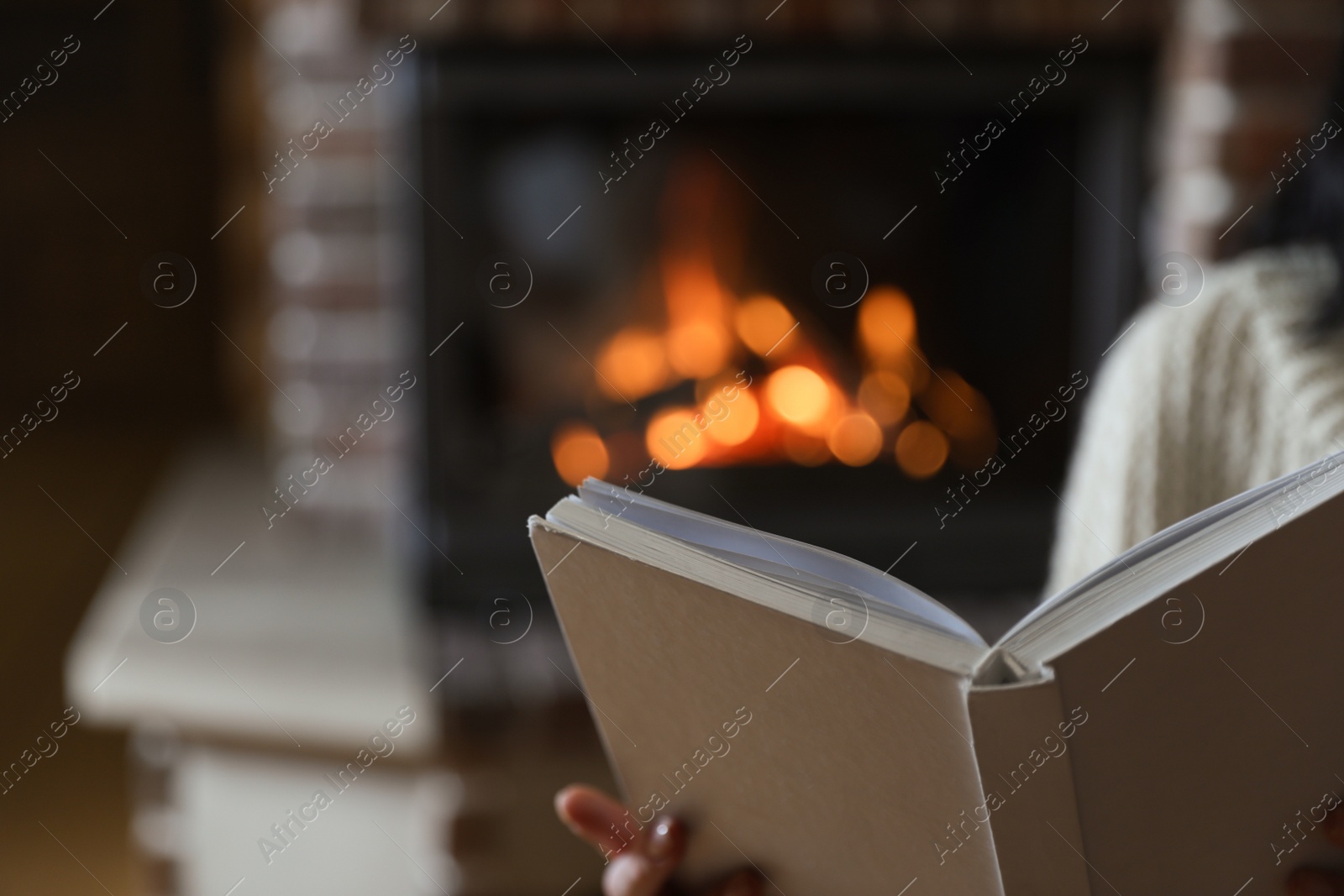 Photo of Woman reading book near burning fireplace at home, closeup