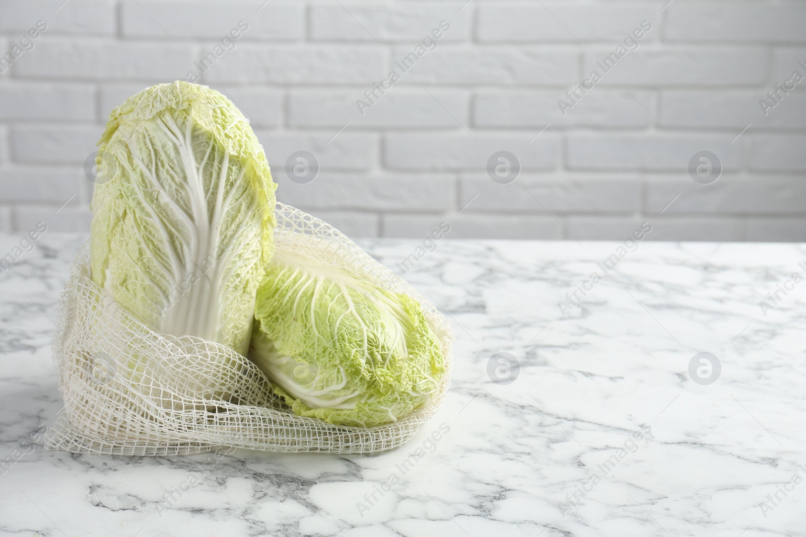 Photo of Fresh Chinese cabbages in string bag on white marble table near brick wall. Space for text