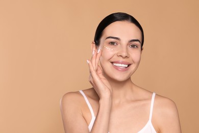 Photo of Beautiful young woman with cream on skin under eye against beige background. Space for text