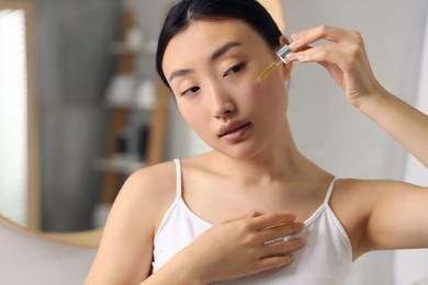 Beautiful young woman applying cosmetic serum onto her face in bathroom, space for text