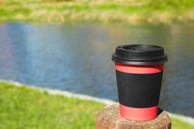 Photo of Hot coffee in takeaway paper cup on wooden stump against river outdoors, space for text