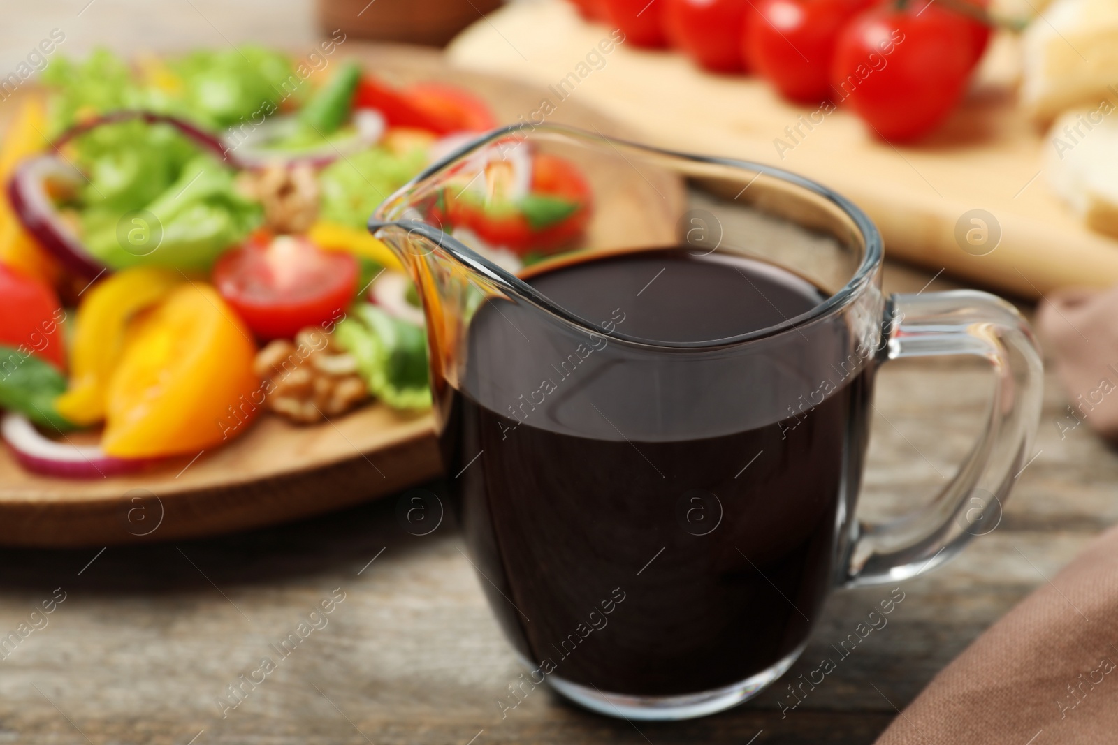 Photo of Balsamic vinegar in glass dish near plate with vegetable salad on wooden table, closeup