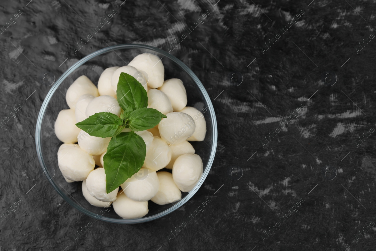 Photo of Tasty mozzarella balls and basil leaves in bowl on black textured table, top view. Space for text
