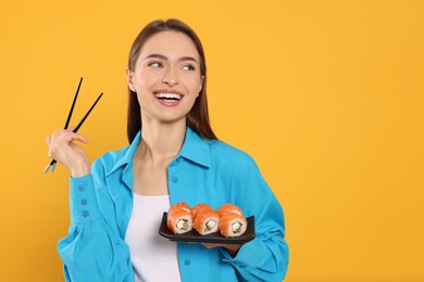 Photo of Happy young woman with plate of sushi rolls and chopsticks on orange background. Space for text