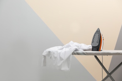 Photo of New modern iron and clean shirt on board against color background, space for text. Laundry day