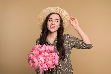 Photo of Beautiful young woman in straw hat with bouquet of pink peonies against light brown background