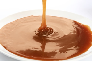 Tasty caramel sauce pouring into plate on white background