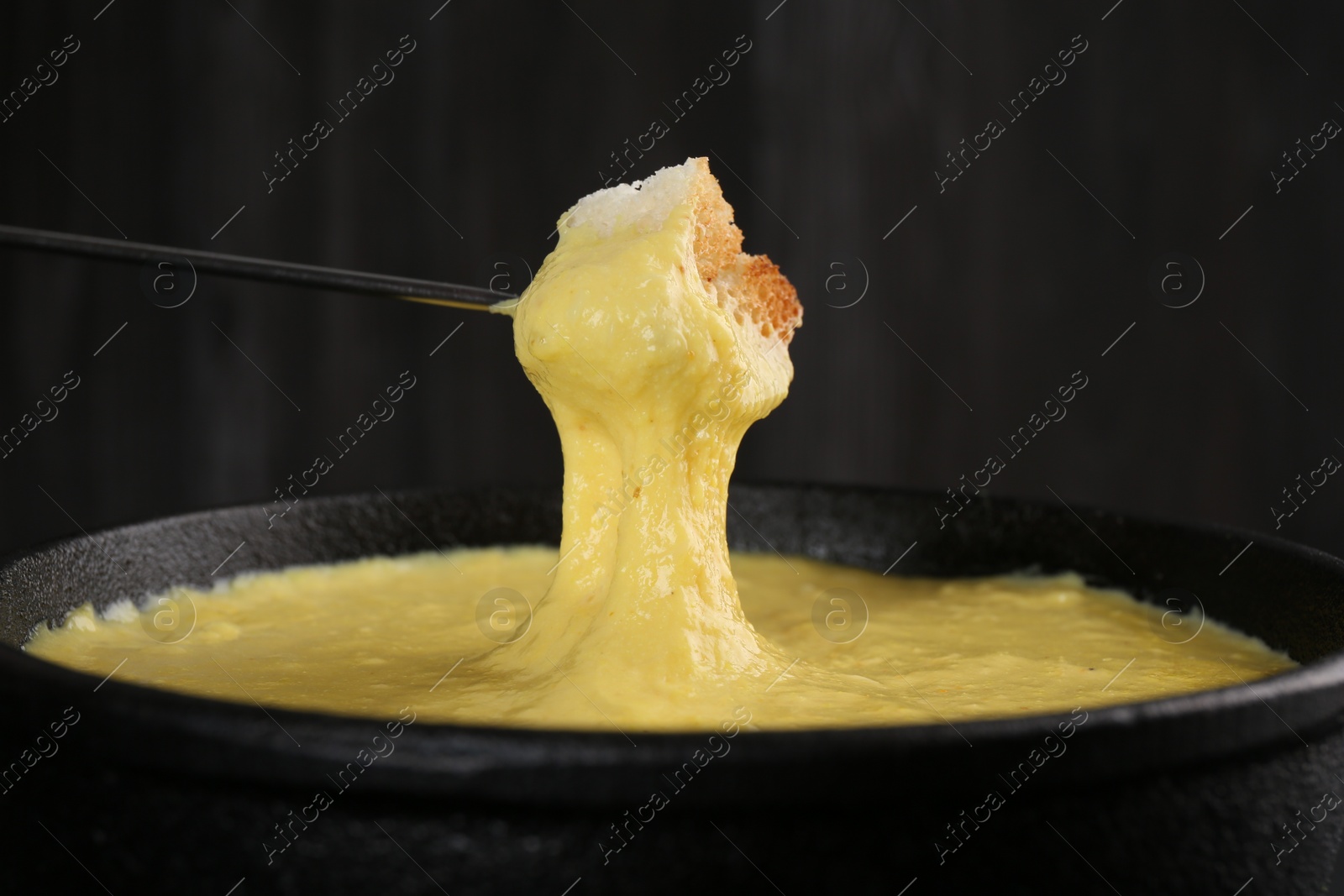 Photo of Dipping piece of bread into fondue pot with tasty melted cheese against dark gray background, closeup