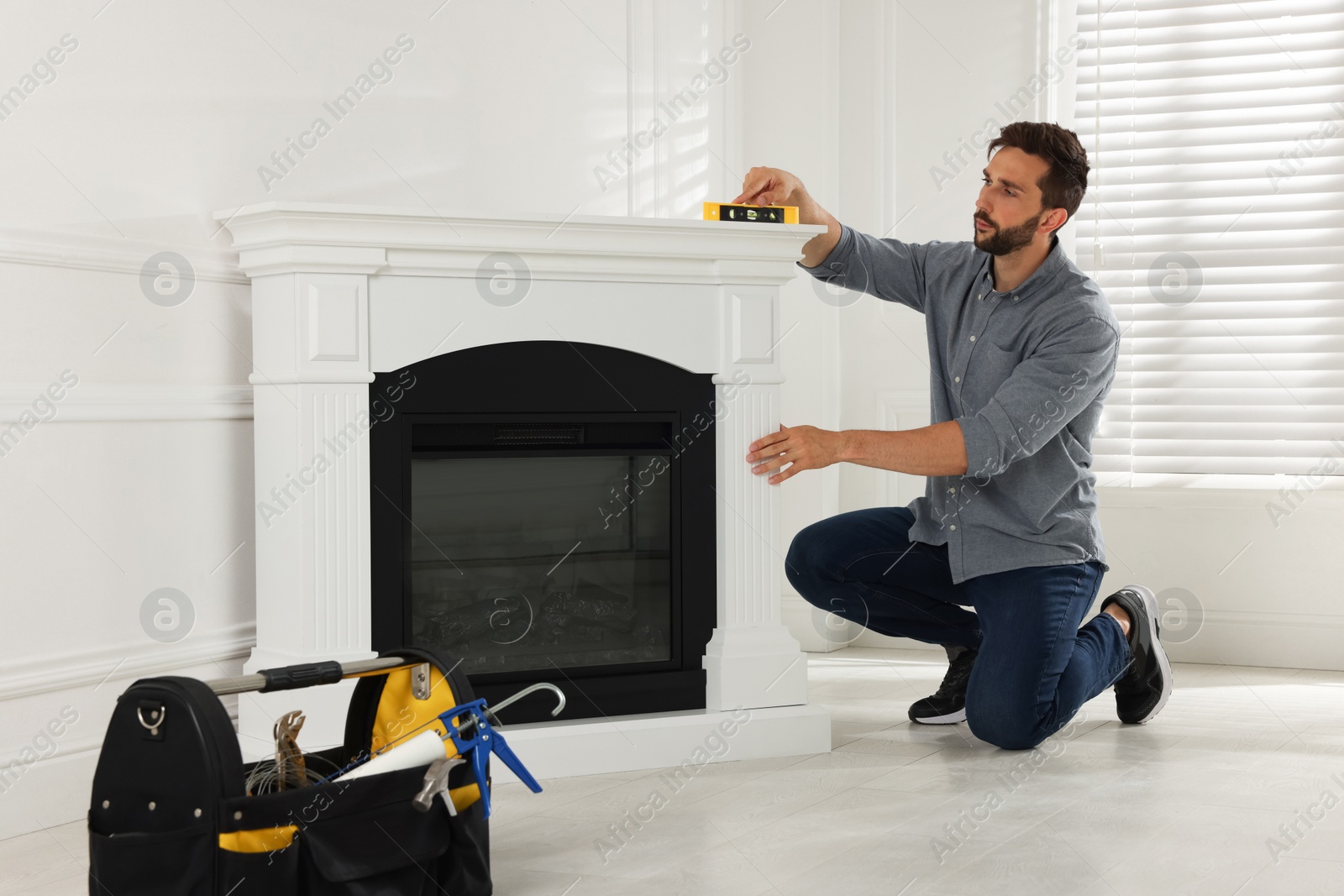 Photo of Man using construction level for installing electric fireplace near white wall in room