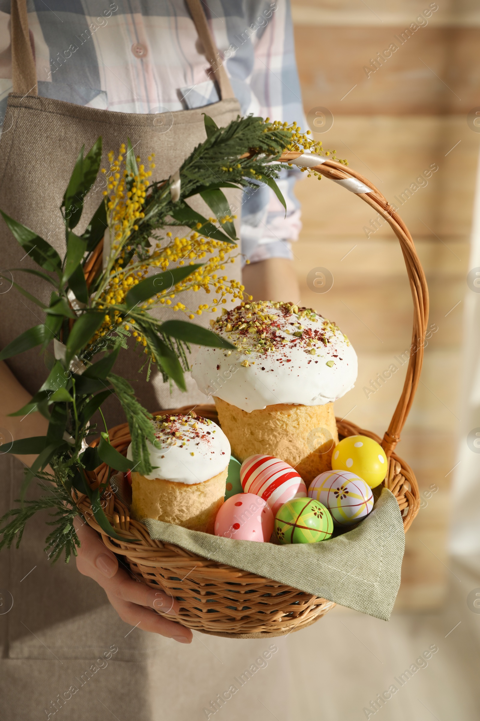 Photo of Woman holding basket with traditional Easter cakes, dyed eggs and flowers indoors, closeup