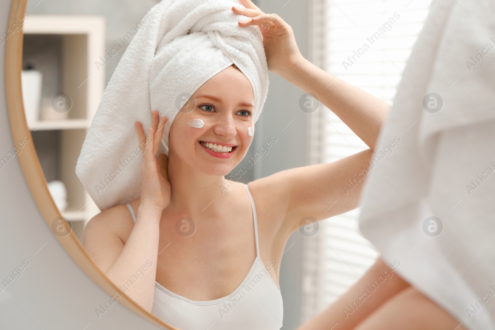 Photo of Smiling woman with freckles and cream on her face near mirror in bathroom