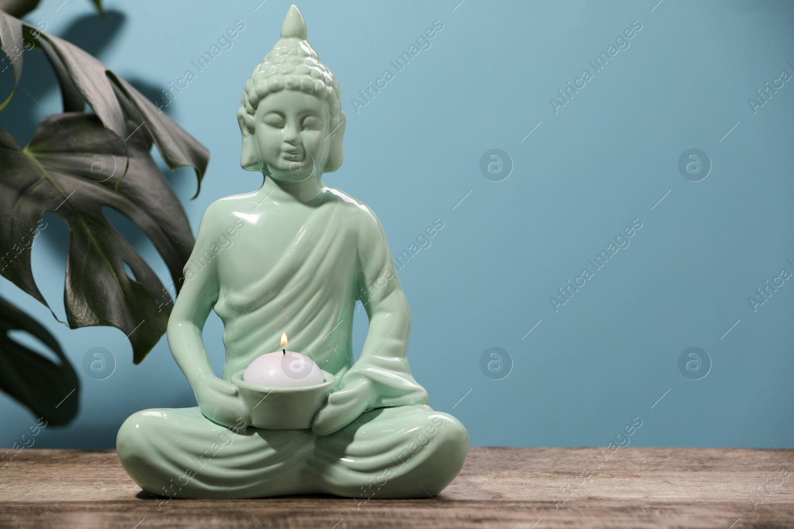Photo of Buddhism religion. Decorative Buddha statue with burning candle on wooden table and monstera against light blue wall, space for text