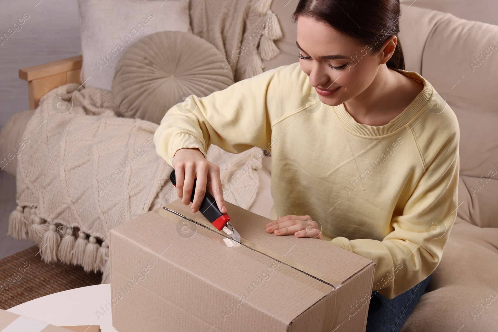 Photo of Young woman using utility knife to open parcel at home