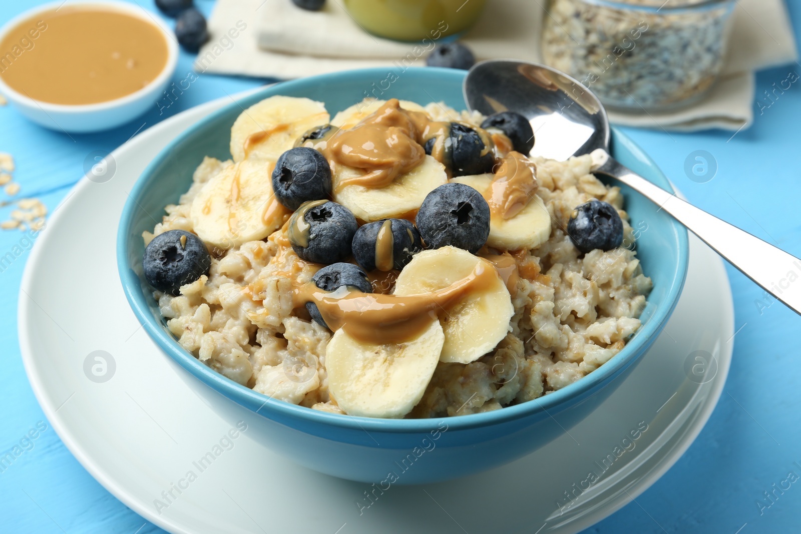 Photo of Tasty oatmeal with banana, blueberries and peanut butter served in bowl on light blue wooden table