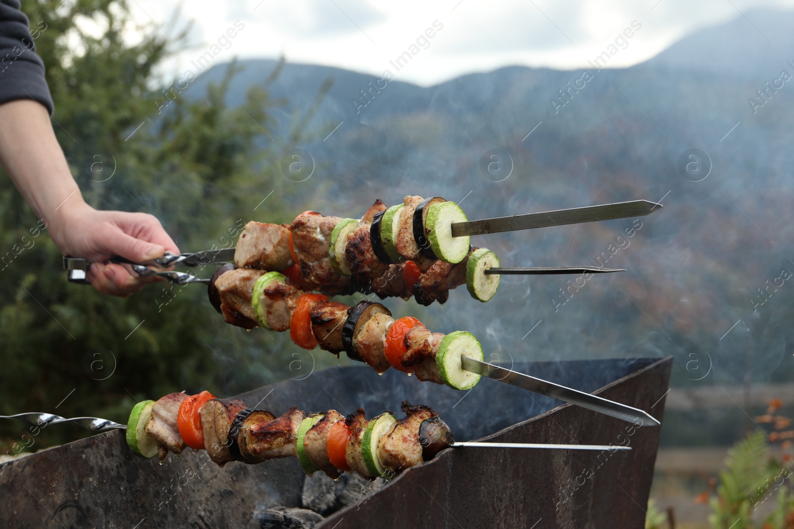 Photo of Woman cooking meat and vegetables on brazier against mountain landscape, closeup