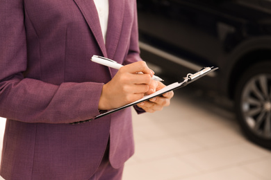 Photo of Young saleswoman with clipboard in modern car salon, closeup