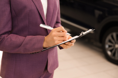 Young saleswoman with clipboard in modern car salon, closeup