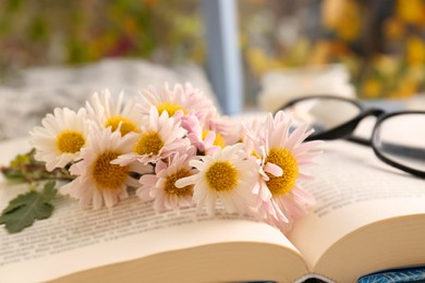 Photo of Open book with chamomile flowers as bookmark and glasses near window, closeup