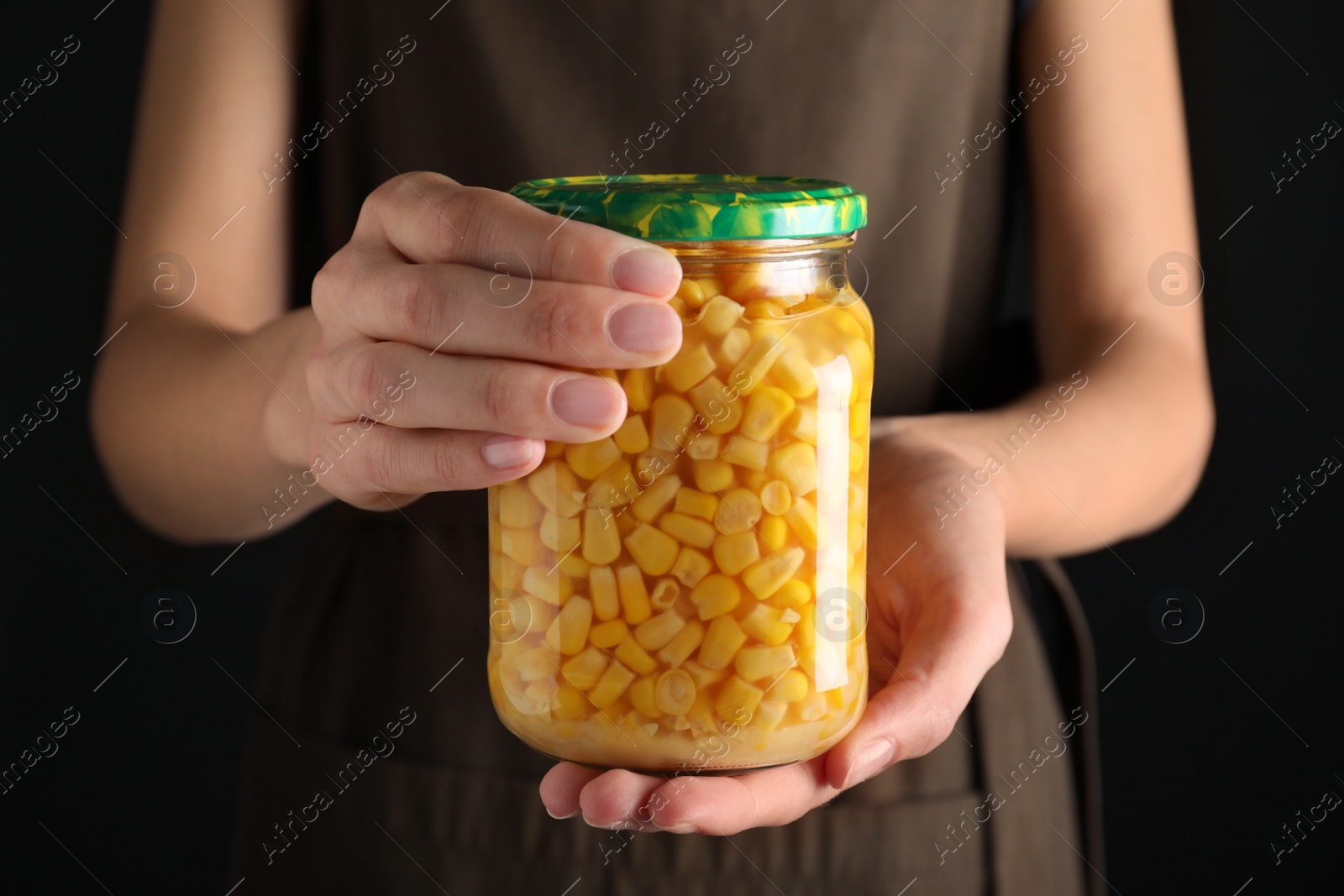 Photo of Woman holding jar with pickled corn against black background, closeup