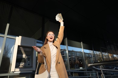 Photo of Excited young woman with money near cash machine outdoors