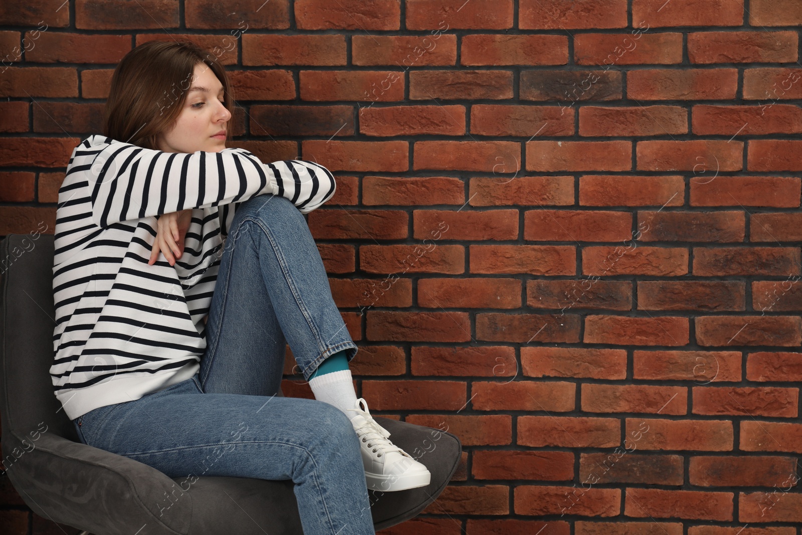 Photo of Sad young woman sitting on chair near brick wall, space for text