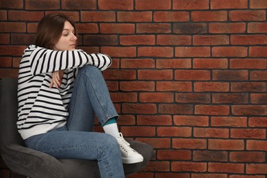 Sad young woman sitting on chair near brick wall, space for text