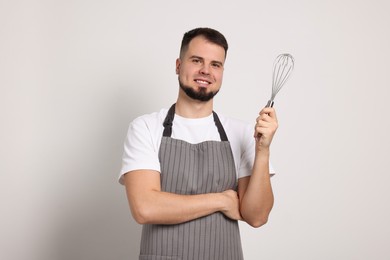 Happy professional confectioner in apron holding whisk on light grey background