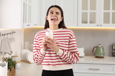 Funny woman with milk mustache holding glass of tasty dairy drink in kitchen