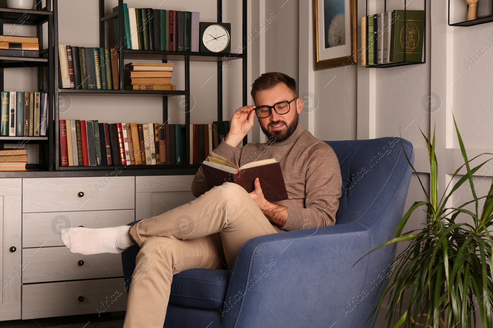 Photo of Young man reading book in armchair indoors. Home library