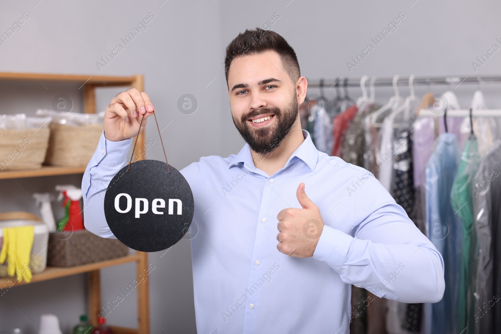 Photo of Dry-cleaning service. Happy worker holding Open sign and showing thumb up indoors