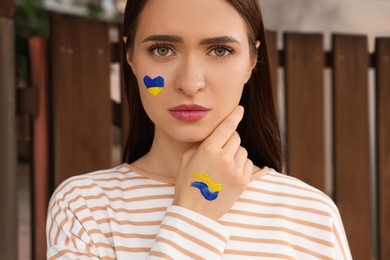 Photo of Young woman with drawings of Ukrainian flag on face outdoors, closeup