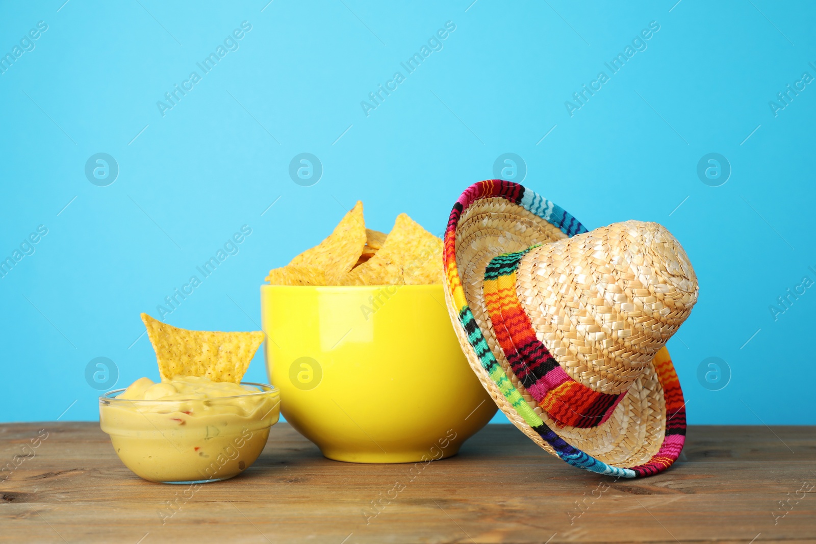 Photo of Mexican sombrero hat, nachos chips and guacamole on wooden table against light blue background