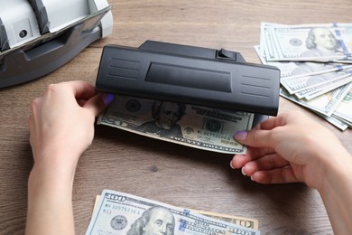 Photo of Woman checking dollar banknote with currency detector at wooden table, closeup. Money examination device