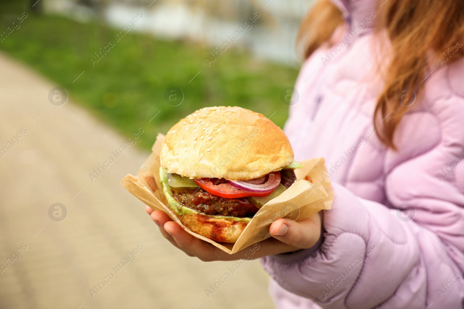 Photo of Little girl holding fresh delicious burger outdoors, closeup. Street food