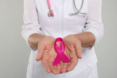 Photo of Doctor with stethoscope holding pink ribbon on light grey background, closeup. Breast cancer awareness