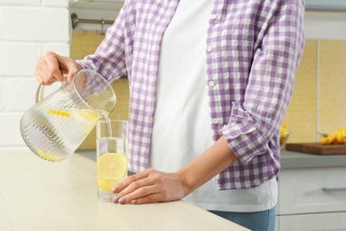 Photo of Young woman pouring lemon water into glass from jug in kitchen, closeup