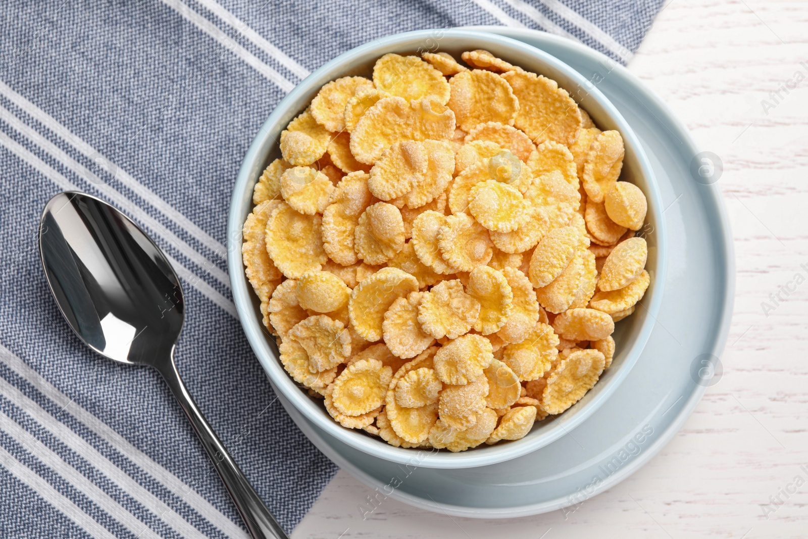 Photo of Bowl of tasty corn flakes and spoon on white wooden table, flat lay