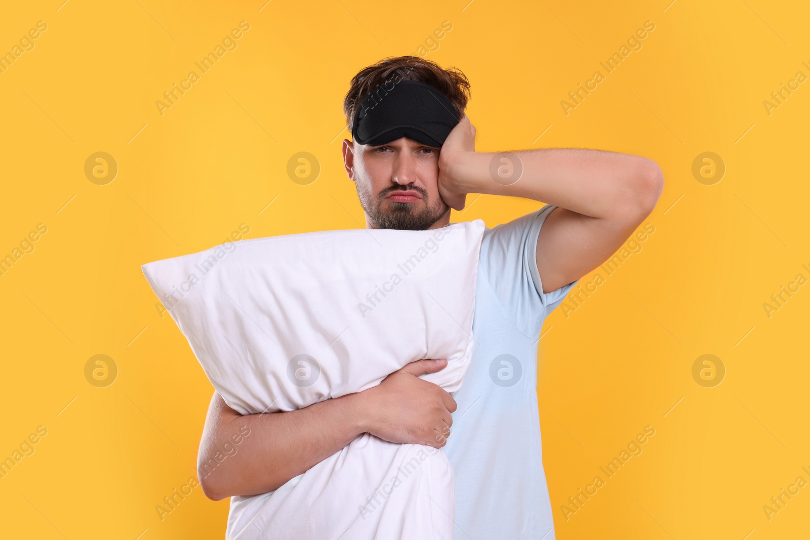 Photo of Unhappy man with pillow and sleep mask on yellow background. Insomnia problem
