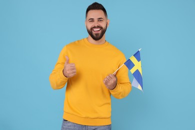 Photo of Young man with flag of Sweden showing thumb up on light blue background