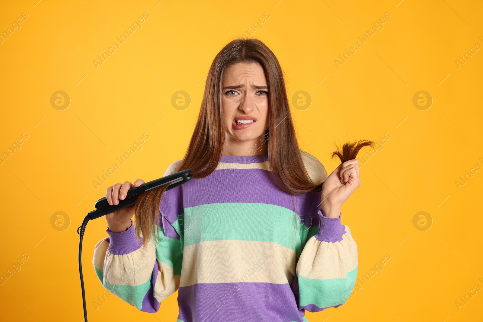 Photo of Upset young woman with flattening iron on yellow background. Hair damage