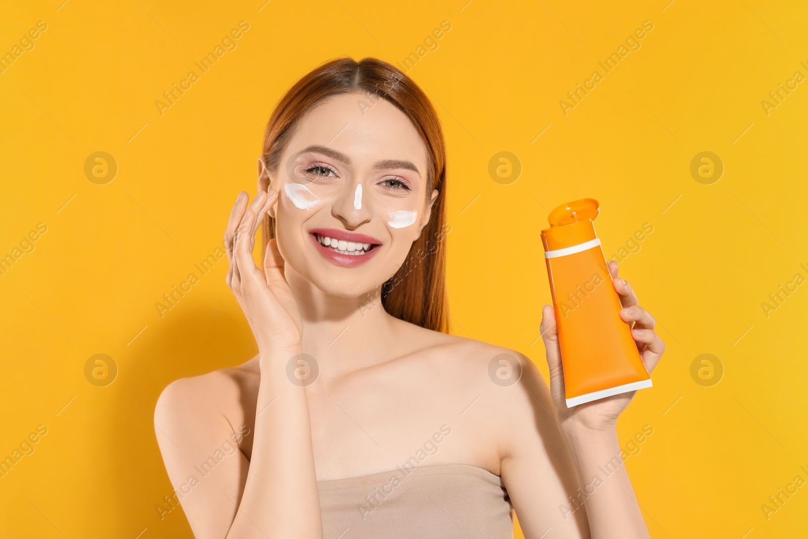 Photo of Beautiful young woman with sun protection cream on her face holding sunscreen against orange background