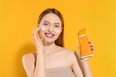 Beautiful young woman with sun protection cream on her face holding sunscreen against orange background