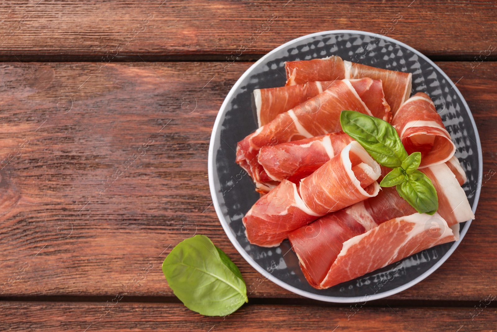 Photo of Plate with rolled slices of delicious jamon and basil on wooden table, top view. Space for text