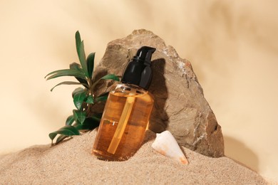 Bottle of serum, stone and green leaves on sand against beige background