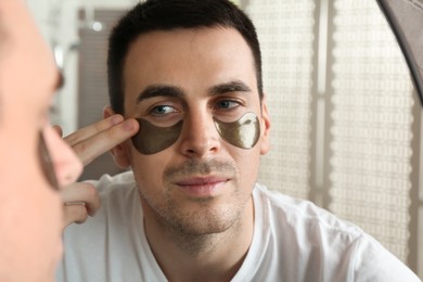 Photo of Young man applying under eye patches near mirror at home, closeup