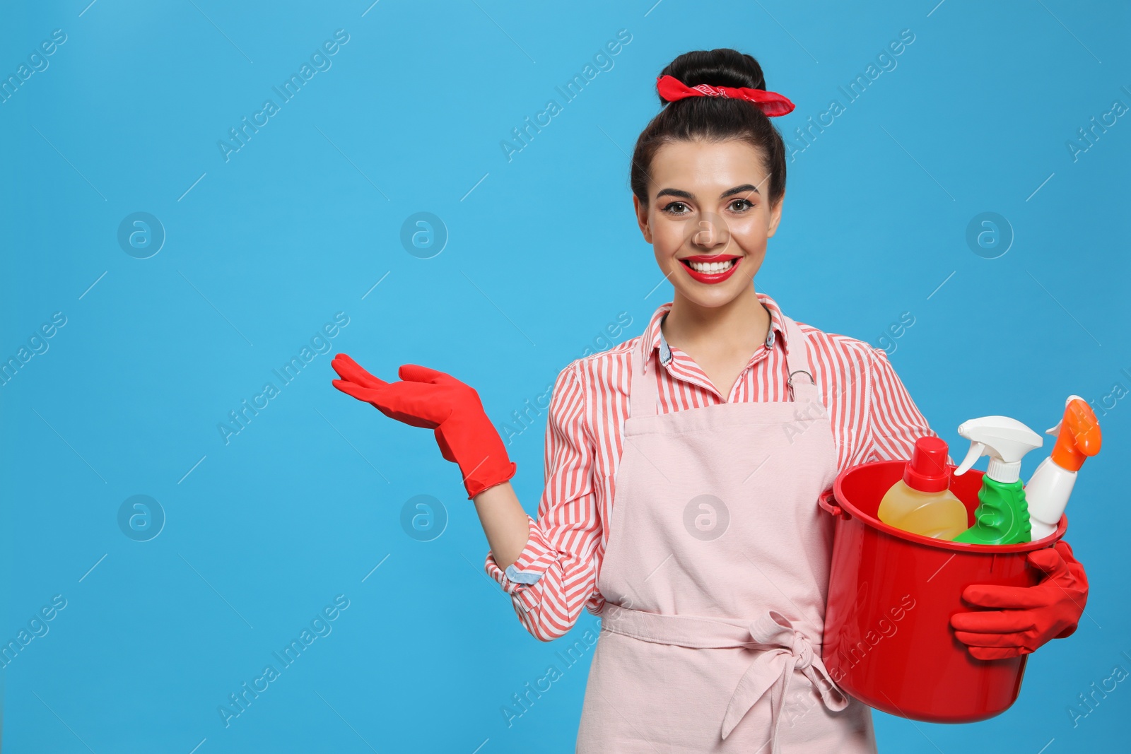 Photo of Young housewife holding bucket with cleaning supplies on light blue background, space for text