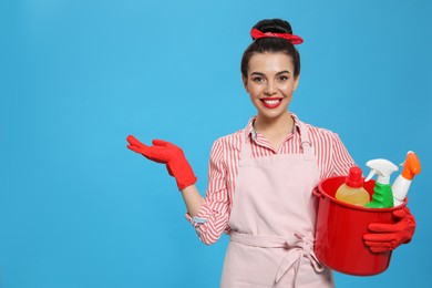 Photo of Young housewife holding bucket with cleaning supplies on light blue background, space for text