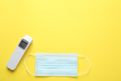 Infrared thermometer and medical mask on yellow background, flat lay with space for text. Checking temperature during Covid-19 pandemic